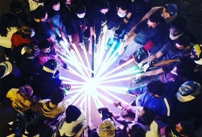 Members of the Jedi Knight Academy Mexico City at night gathered in a circle with each person extending their incandescent lightsabers to touch all together at the circle's center.
